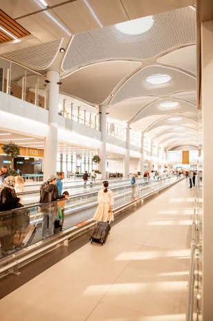 A spacious modern airport terminal with a high, curved ceiling featuring circular lights. Several travelers walk along a moving walkway, some pulling suitcases. The area is brightly lit with natural light from large windows, creating a clean and airy atmosphere. Shops and gates are visible in the background.