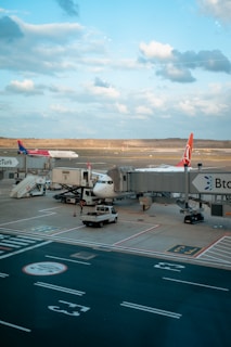 Airplanes are parked at an airport gate, with one being serviced by various vehicles and equipment. It is a clear day with a blue sky and scattered clouds. The tarmac and runway are visible, along with ground vehicles and jet bridges connecting to the planes.