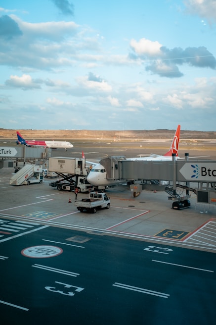 Airplanes are parked at an airport gate, with one being serviced by various vehicles and equipment. It is a clear day with a blue sky and scattered clouds. The tarmac and runway are visible, along with ground vehicles and jet bridges connecting to the planes.