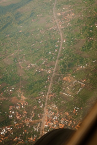 An aerial view of a rural area with scattered buildings and dense green vegetation. A road runs vertically through the center of the image, surrounded by patches of farmland and small clusters of houses. The image is taken from above, possibly from an aircraft, as part of the aircraft frame is visible in the bottom right corner.