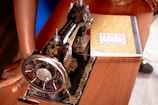 A vintage sewing machine with intricate gold embellishments is situated on a wooden table. Beside it lies a yellow composition notebook. Partial view of a person's hand resting on the table gives a sense of presence and action.