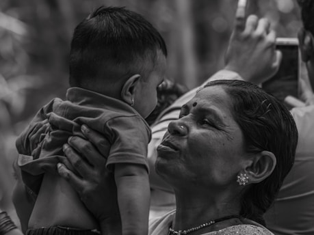 A woman holds a baby up closely, appearing to engage with the child in an affectionate manner. The setting seems to have other people, one of whom is taking a photo or video in the background. The image is in black and white, highlighting the textures and emotions on the woman's face.