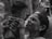 A woman holds a baby up closely, appearing to engage with the child in an affectionate manner. The setting seems to have other people, one of whom is taking a photo or video in the background. The image is in black and white, highlighting the textures and emotions on the woman's face.