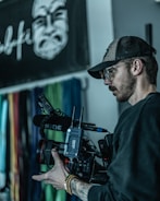A person is holding a professional video camera, with a microphone attached. They are wearing a baseball cap, glasses, and a dark shirt. In the background, there is a blurred wall with colorful vertical lines and a black and white graphic of a face.