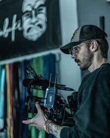 A person is holding a professional video camera, with a microphone attached. They are wearing a baseball cap, glasses, and a dark shirt. In the background, there is a blurred wall with colorful vertical lines and a black and white graphic of a face.