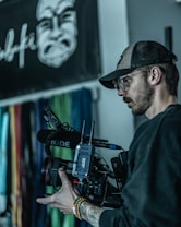 A person is holding a professional video camera, with a microphone attached. They are wearing a baseball cap, glasses, and a dark shirt. In the background, there is a blurred wall with colorful vertical lines and a black and white graphic of a face.