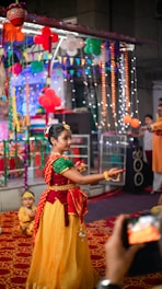 A girl in traditional Indian attire is dancing in a festive environment decorated with colorful balloons, lights, and banners. Other people are present in the background, observing the performance. A child in a similar costume sits on the floor nearby, and a phone is capturing the event.