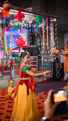 A girl in traditional Indian attire is dancing in a festive environment decorated with colorful balloons, lights, and banners. Other people are present in the background, observing the performance. A child in a similar costume sits on the floor nearby, and a phone is capturing the event.