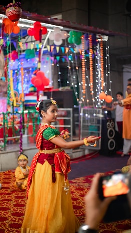A girl in traditional Indian attire is dancing in a festive environment decorated with colorful balloons, lights, and banners. Other people are present in the background, observing the performance. A child in a similar costume sits on the floor nearby, and a phone is capturing the event.