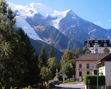 A picturesque mountain landscape featuring snow-capped peaks under a clear blue sky. A quaint village with traditional European architecture is nestled at the base of the mountains, surrounded by lush green trees. Some buildings are adorned with flags, and there are street signs and vehicles visible on the road.