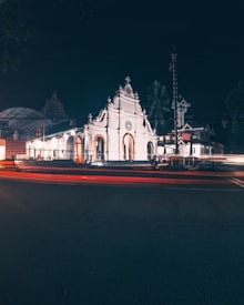 A brightly illuminated white church set against a dark night sky. The church features intricate architectural details, including arched windows and ornate carvings. Light trails from passing vehicles add dynamic streaks of red and orange across the foreground, while nearby trees are faintly visible in the background.