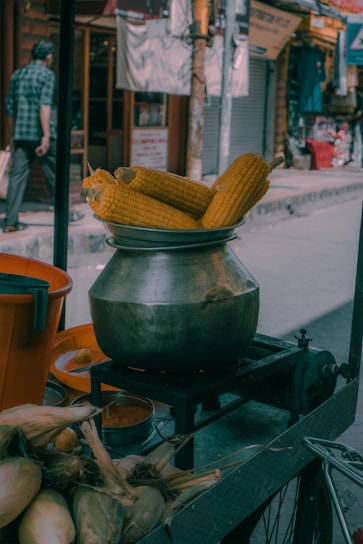 A street scene displaying a metal pot filled with corn cobs atop a stove, set in an urban environment with a passerby visible in the background. Corn husks and a bucket are also part of the setup, indicating a street food setting.