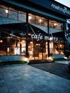 A modern cafe with large glass windows displaying a warm and inviting interior with visible lights. The building has a stylish design with plants by the entrance, and the name 'cafe metro' is displayed prominently. There are outdoor lighting fixtures casting a soft glow on the facade.