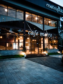A modern cafe with large glass windows displaying a warm and inviting interior with visible lights. The building has a stylish design with plants by the entrance, and the name 'cafe metro' is displayed prominently. There are outdoor lighting fixtures casting a soft glow on the facade.