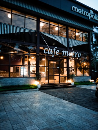 A modern cafe with large glass windows displaying a warm and inviting interior with visible lights. The building has a stylish design with plants by the entrance, and the name 'cafe metro' is displayed prominently. There are outdoor lighting fixtures casting a soft glow on the facade.