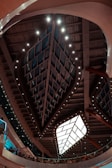 An architectural interior featuring a large geometric ceiling structure with a central illuminated panel surrounded by patterned glass. The ceiling is adorned with rows of lights, creating an intricate design. Below the ceiling, people are gathered along a curved balcony, and the walls have modern glass panels with a mixture of warm and cool lighting tones.