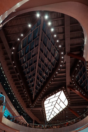 An architectural interior featuring a large geometric ceiling structure with a central illuminated panel surrounded by patterned glass. The ceiling is adorned with rows of lights, creating an intricate design. Below the ceiling, people are gathered along a curved balcony, and the walls have modern glass panels with a mixture of warm and cool lighting tones.