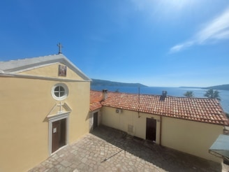 A small chapel with a cross on its roof stands next to a building with a terracotta-tiled roof. The buildings are situated on a stone courtyard overlooking a calm, blue sea with distant hills under a clear sky.