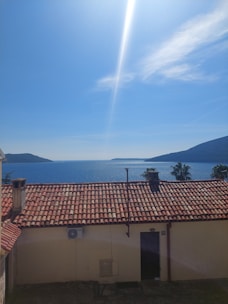 A rooftop with red tiles overlooks a calm sea, with two distant landmasses in the background beneath a clear blue sky. A chimney and an air conditioning unit are visible on the roof, and palm trees are seen nearby.