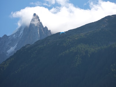A striking mountain peak rises dramatically, partly shrouded by clouds, with a paragliding individual soaring in the clear blue sky. The foreground features a dense forest covering the hillside, contributing to the sense of adventure and natural beauty.