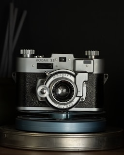 A vintage Kodak 35mm film camera is placed centrally, with metallic and textured black surfaces highlighted under low light. The camera's intricate dials and lens details are visible, with a focus on its classic design. It sits atop a couple of stacked film reels.