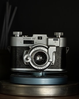 A vintage Kodak 35mm film camera is placed centrally, with metallic and textured black surfaces highlighted under low light. The camera's intricate dials and lens details are visible, with a focus on its classic design. It sits atop a couple of stacked film reels.