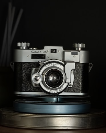 A vintage Kodak 35mm film camera is placed centrally, with metallic and textured black surfaces highlighted under low light. The camera's intricate dials and lens details are visible, with a focus on its classic design. It sits atop a couple of stacked film reels.