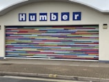 A decorative garage door with horizontal stripes in a variety of bright colors. Above the door is a sign with blue letters reading 'Humber'. The building facade is light-colored and the ground is paved.