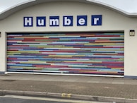 A decorative garage door with horizontal stripes in a variety of bright colors. Above the door is a sign with blue letters reading 'Humber'. The building facade is light-colored and the ground is paved.