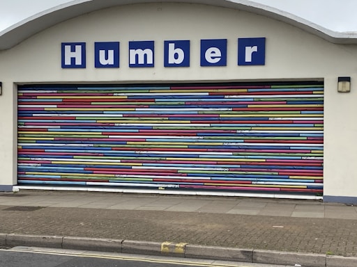 A decorative garage door with horizontal stripes in a variety of bright colors. Above the door is a sign with blue letters reading 'Humber'. The building facade is light-colored and the ground is paved.