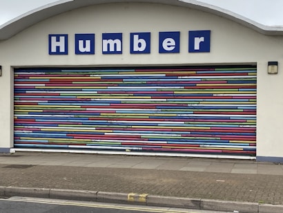 A decorative garage door with horizontal stripes in a variety of bright colors. Above the door is a sign with blue letters reading 'Humber'. The building facade is light-colored and the ground is paved.