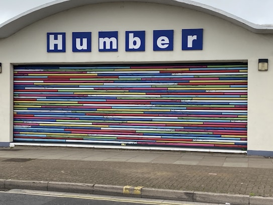 A decorative garage door with horizontal stripes in a variety of bright colors. Above the door is a sign with blue letters reading 'Humber'. The building facade is light-colored and the ground is paved.