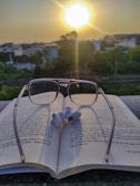 A pair of reading glasses placed on an open book with text in Hindi script. Earbuds rest on top of the book. In the background, the sun is setting, casting a warm glow over a landscape of trees and buildings.