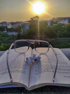 A pair of reading glasses placed on an open book with text in Hindi script. Earbuds rest on top of the book. In the background, the sun is setting, casting a warm glow over a landscape of trees and buildings.