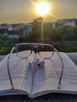 A pair of reading glasses placed on an open book with text in Hindi script. Earbuds rest on top of the book. In the background, the sun is setting, casting a warm glow over a landscape of trees and buildings.