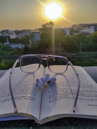 A pair of reading glasses placed on an open book with text in Hindi script. Earbuds rest on top of the book. In the background, the sun is setting, casting a warm glow over a landscape of trees and buildings.
