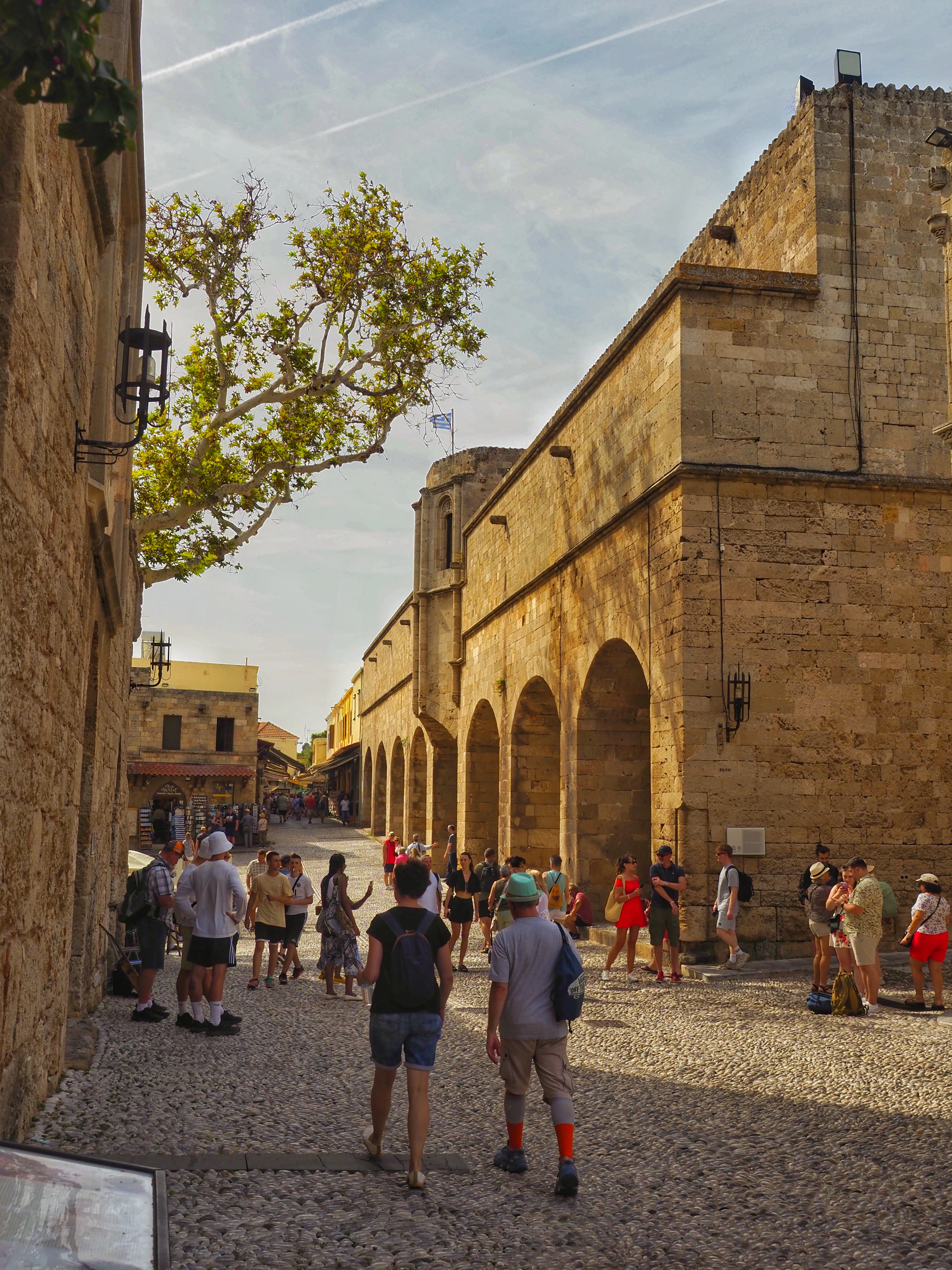 A sunlit medieval street inside Rhodes Old Town, with tourists walking past stone arches and a shade tree.