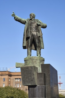A large bronze statue of a man standing atop a pedestal, with one arm outstretched in a gesture of leadership or speech. The background includes a clear blue sky, a building with classical architecture, and some trees. The statue is highly detailed, capturing the man's clothing in a realistic manner.