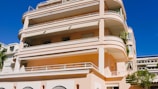A large, modern apartment building with multiple balconies and terraces. The structure features smooth, rounded edges and is painted in a pale pink color. It has several floors and large windows, with some greenery visible on the terraces.