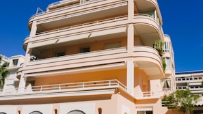 A large, modern apartment building with multiple balconies and terraces. The structure features smooth, rounded edges and is painted in a pale pink color. It has several floors and large windows, with some greenery visible on the terraces.