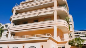 A large, modern apartment building with multiple balconies and terraces. The structure features smooth, rounded edges and is painted in a pale pink color. It has several floors and large windows, with some greenery visible on the terraces.