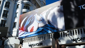 A large billboard on the side of a building features a high-fashion advertisement. The image on the billboard shows a person blindfolded with a silky fabric, wearing large earrings and bright red lipstick. The building in the background has an ornate architectural style with several windows.