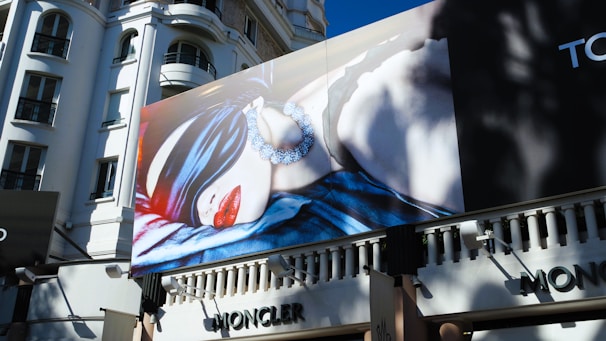 A large billboard on the side of a building features a high-fashion advertisement. The image on the billboard shows a person blindfolded with a silky fabric, wearing large earrings and bright red lipstick. The building in the background has an ornate architectural style with several windows.