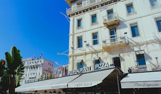 An elegant urban scene featuring a classic European-style building with ornate balconies and windows. The facade is adorned with cafe and restaurant signage reading 'Caffe Roma' and 'Restaurant Pizza'. The sky is bright blue, suggesting a clear, sunny day, and palm trees are visible at the edge of the scene.