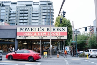 A busy urban street corner with a bookstore named Powell's Books. The large sign advertises used and new books along with book event dates. Several pedestrians are walking by, and cars are driving past, including a noticeable red sedan. A tall modern apartment building forms the backdrop, alongside green trees and streetlights on the sidewalk.