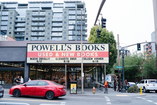 A busy urban street corner with a bookstore named Powell's Books. The large sign advertises used and new books along with book event dates. Several pedestrians are walking by, and cars are driving past, including a noticeable red sedan. A tall modern apartment building forms the backdrop, alongside green trees and streetlights on the sidewalk.