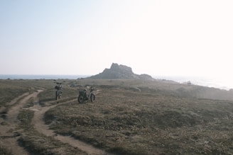 Two motorcycles are parked on a dirt path leading through a grassy terrain towards a rocky outcrop near the sea. The scene is bathed in soft light, suggesting either early morning or late afternoon. The ocean is visible in the background, and the atmosphere is calm and peaceful.