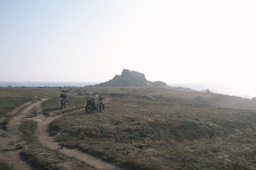 Two motorcycles are parked on a dirt path leading through a grassy terrain towards a rocky outcrop near the sea. The scene is bathed in soft light, suggesting either early morning or late afternoon. The ocean is visible in the background, and the atmosphere is calm and peaceful.