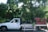 A white pickup truck loaded with a variety of potted plants, including small trees and shrubs, is parked on a street. The truck bed is partially framed with wooden planks painted in red. The lush greenery contrasts with the vehicle, set against a backdrop of tall trees and shrubs under bright daylight.