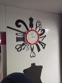 A wall clock with a red frame is positioned in the center of the image. Surrounding the clock are various black silhouettes of sewing tools, including scissors, a dress form, a ruler, and a spool of thread. Below the clock is a black mannequin torso. The background is a white, tiled wall.